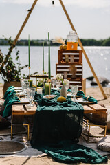 Picnic on beach at sunset in rustic style, boho conception. Decor with blanket, pillows, arch, garlands, wooden decor, candles, flowers, dry leaves, straw chair. Bachelorette party, birthday, wedding.