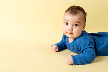 Naklejka premium A beautiful little baby in a blue bodysuit, lies on the stomach on a yellow background. Concept, mockup, blank, copy space.