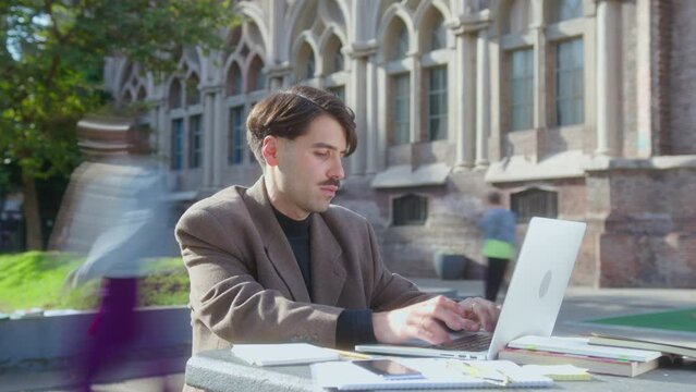 Time lapse of young man with mustache in smart casual outfit sitting outdoors on university campus and typing on laptop, students walking in the background