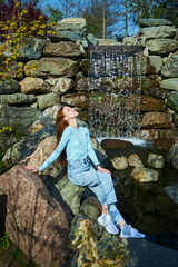 A stylish beautiful young girl is sitting on a rock by a waterfall. Photo shoot in the park