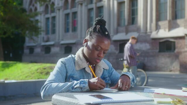 Time lapse of young smart African-American girl in wireless earbuds sitting outdoors on campus, doing homework and writing down in workbook, students walking in the background