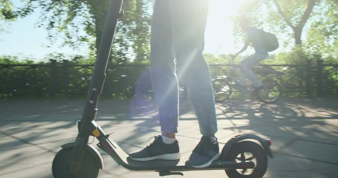 Woman on electric scooter overtakes man on bicycle. City park, embankment, summer weather, bright sun.