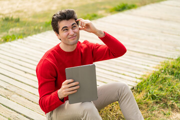 Fototapeta premium Young handsome man holding a tablet at outdoors having doubts and with confuse face expression