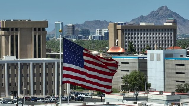 American flag with Phoenix, Arizona state capitol and government buildings in background. Aerial establishing shot with long zoom lens.