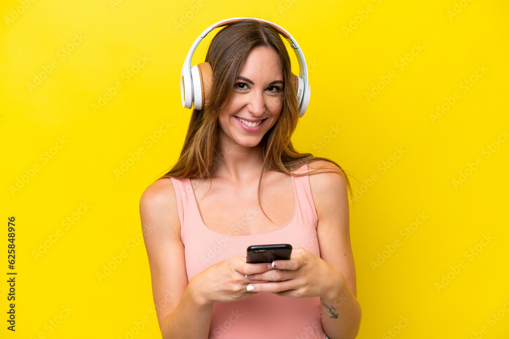 Wall mural Young caucasian woman isolated on yellow background listening music with a mobile and looking front