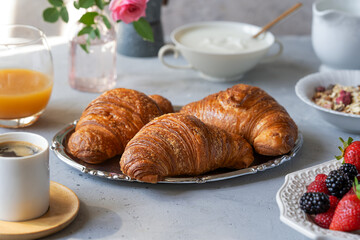 Breakfast in the morning with fresh baked croissants, berries, muesli, coffee, milk, yogurt, honey and juice over grey background