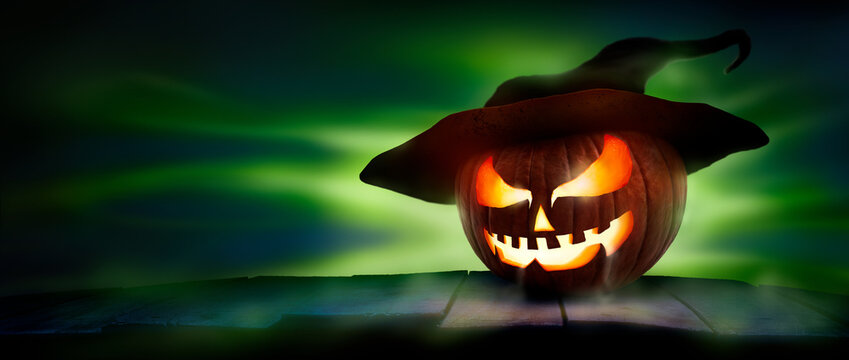 A spooky halloween pumpkin, Jack O Lantern, with an evil spinning green aura glow on a wooden table against a dark background with a crescent moon and tree silhouette.
