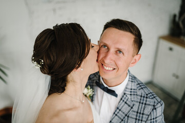 Bride kisses in cheek groom at home. First meeting is newlyweds in interior in bedroom. Couple in love.  Handsome man and woman posing together. Happy wedding day of marriage. Closeup top view