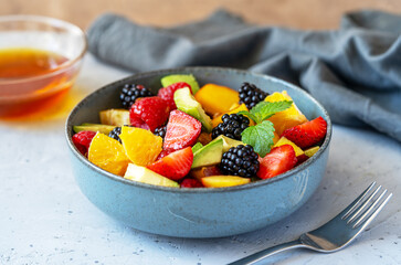 Healthy fruit salad in a grey bowl made with avocado, strawberry, raspberry, orange, blackberry, almonds and peaches. Yogurt, honey behind. Grey background