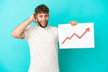 Young handsome caucasian man isolated on blue background holding a sign with a growing statistics arrow symbol with surprised expression