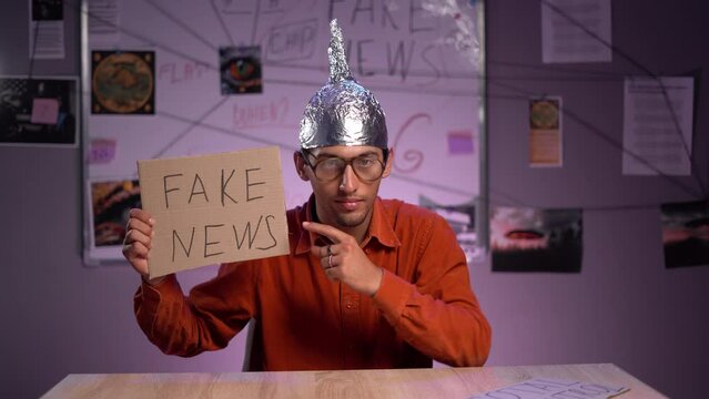 Young conspiracy theorist in aluminum cap is sitting in his office and showing fake news. Conspiracy theory concept