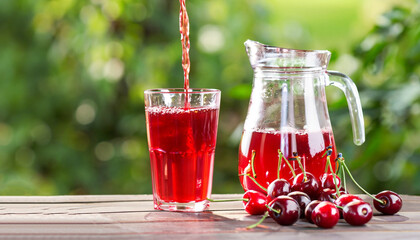 cherry juice pouring from jug into glass and ripe berries on wooden table outdoors. Summer refreshing drink