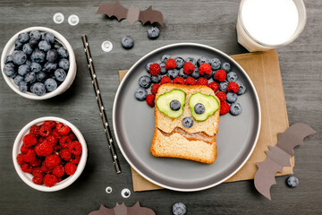 Funny scary monster face smile on halloween sandwich toast bread with peanut butter, blueberry, raspberry on plate,bats on gray background close up. Kids child sweet dessert breakfast lunch food