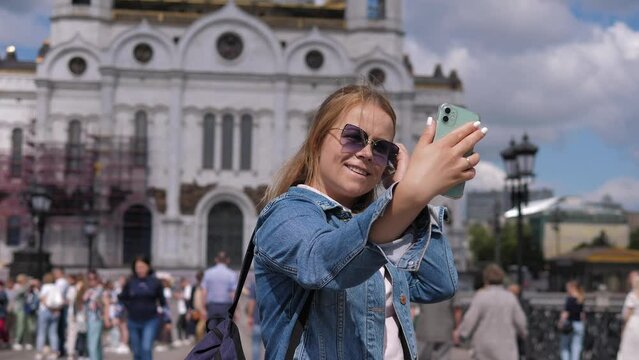 Portrait of a young woman who shoots a video standing against the background of a church and a lot of people. A tourist takes a selfie on her phone with the city during a trip.