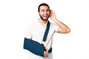 Young handsome man with broken arm and wearing a sling over isolated chroma key background listening to something by putting hand on the ear