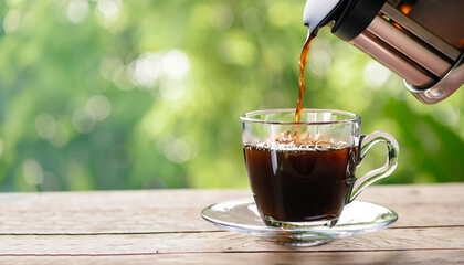 closeup coffee pouring from french press into glass cup on wooden table with green blurred...