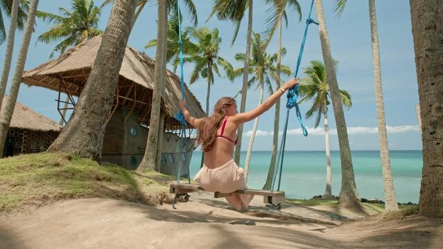Summer of young woman sitting on swing on the beach on blue sea and green palms background and having fun. Happy female relaxing in hammock on a tropical beach. Young girls traveler enjoying. Summer
