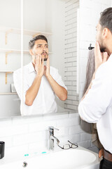 Handsome businessman looking at mirror and applying moisturizing cream on cheeks in bathroom. Groomed young guy doing skincare morning routine after taking a shower