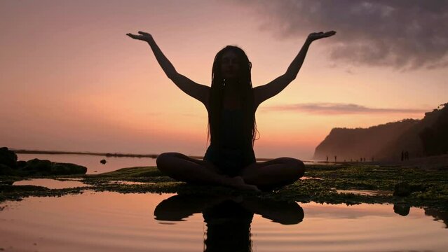 Silhouette of Woman Raising His Hands or Open arms when sun rising up. A young slim mindful female does yoga by the sea. Peaceful girl sit on lotus pose with her raising hands up to greet the dawn