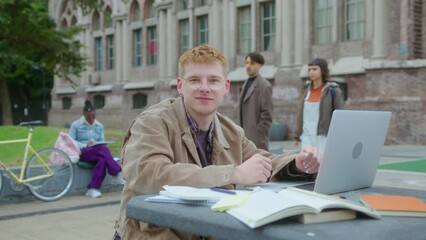 Young guy with earrings and painted nails wearing casual outfit posing for camera with smile, sitting with copybooks and laptop at outdoor table in park-like setting on campus. Medium shot