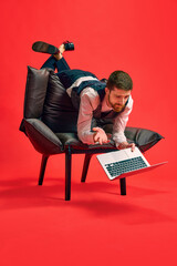 Finishing tasks. Businessman lying on armchair in weird pose, working on laptop against red studio background. Concept of business, working routine, deadlines, freelance, office, ad