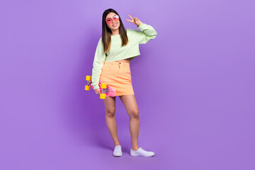 Portrait of friendly optimistic nice girl wear stylish pullover skirt hold skateboard showing v-sign isolated on violet color background
