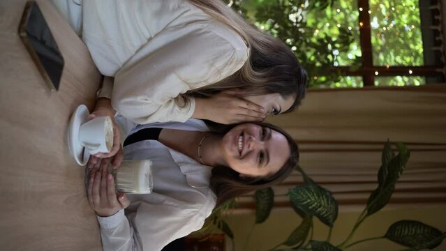Two smiling young women drinking coffee or tea and gossiping at indoor cafe. People communication and friendship concept