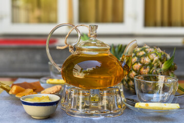 Glass tea pot with pineapple tea and homeade jam,cups and crystalized fruits on dark background