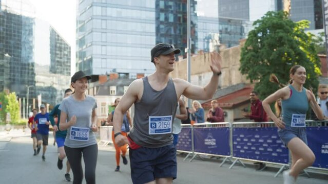 Slow Motion Portrait of Smiling Middle Aged Man Running in a City Marathon, Waving at the Supportive Audience. Friendly Happy Male Runner Celebrating Crossing the Finish Line in a Race