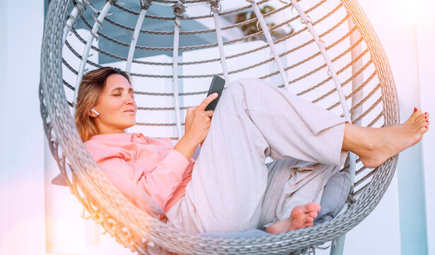 Beautiful middle-aged woman listening Earbuds smartphone music lying in comfortable hanging chair on open house terrace and enjoying the sunset sun rays. Modern technology and home lifestyle concept