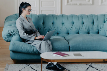 Obraz premium Busy female freelancer on comfortable sofa, dressed formally, multitasks on laptop, project work, and video chats with employee. Remote job concept.