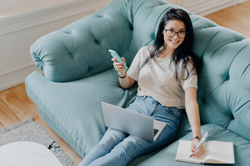 Cheerful female freelancer in casual outfit, writes in notepad, uses laptop, on couch. Student prepares project work at home.