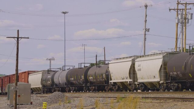 a train comprised of box cars, tanker cars  and hopper cars moves across the railroad tracks surrounded by utility poles