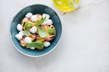 Salad with grilled peaches, mini mozzarella and green basil, flat lay on a light-beige marble background, horizontal shot with space