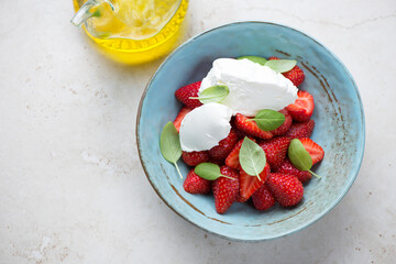 Fresh sliced strawberries served with mascarpone and green basil in a turquoise bowl, elevated view on a light-beige stone background with space
