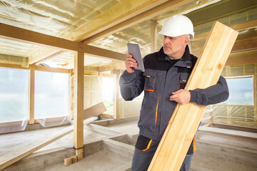 Man builder. Worker with whiteboard and tablet. Guy builder in gray uniform. Brigadier in building house. Construction of wooden buildings. Builder at work. Man in unfinished wooden house