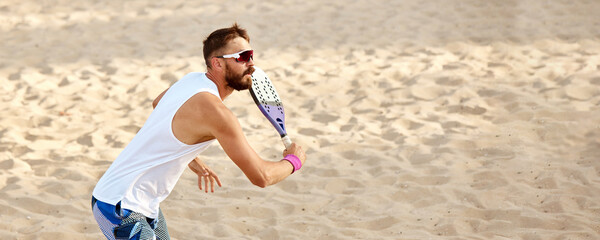 Dynamic image of young man playing beach tennis, hitting ball with racket. Outdoor training on warm...
