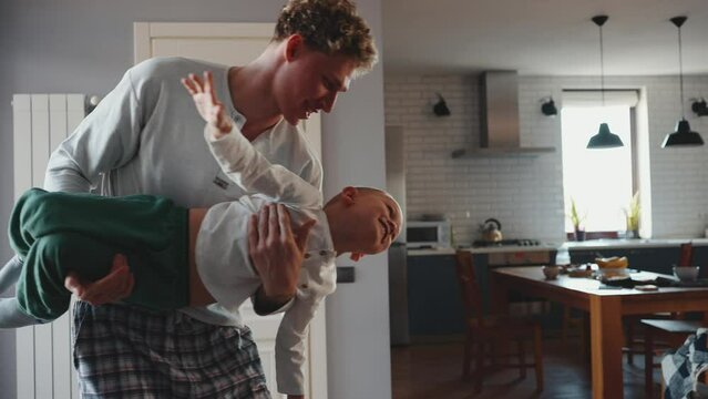 Cheerful curly haired blond father playing while holding his son in his arms depicting airplane at home 