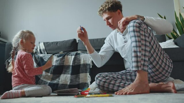 Cute curly haired blond father and daughter drawing in coloring book and talking at home on the floor