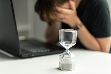 Hourglass in the foreground. A woman is sitting in front of a laptop and is stressed, covering her face with her hands. The concept of lack of time.