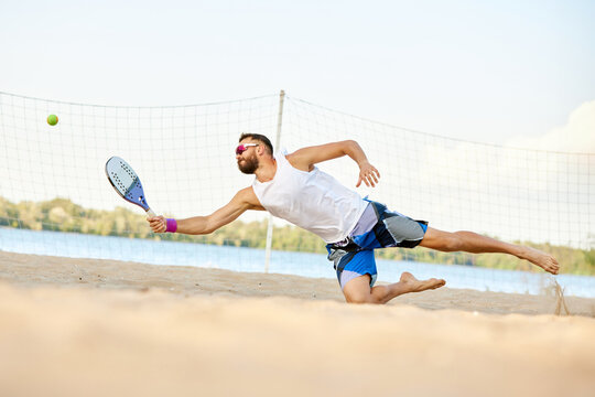 Dynamic image of young man playing beach, paddle tennis, hitting ball with racket and falling down. Outdoor training on warm summer day. Concept of sport, leisure time, active lifestyle, hobby, ad
