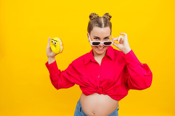 Cheerful pregnant woman holding a sweet yellow donut in her hand on a yellow background. Expecting...