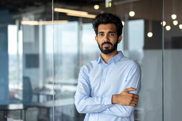 Portrait of a young successful Indian male programmer, developer standing in an office center, crossing his arms over his chest and looking seriously at the camera