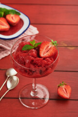 Strawberry granita or fresh berry sorbet in glass on old red wooden table background. Texture of ice cream or sorbet. Ice cream with strawberry and mint. Sorbet recipe. Summer treat. Selective focus.