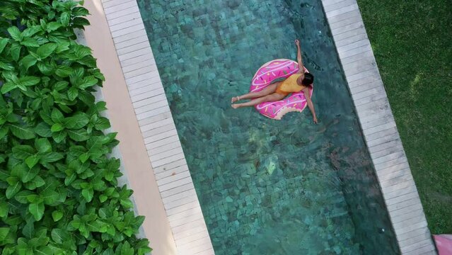 Top view of young woman in swimsuit on the pink donut lilo in the swimming pool. Female relaxing on inflatable float the pool at villa. Inflatable ring and mattress. Summer holiday idyllic. High view