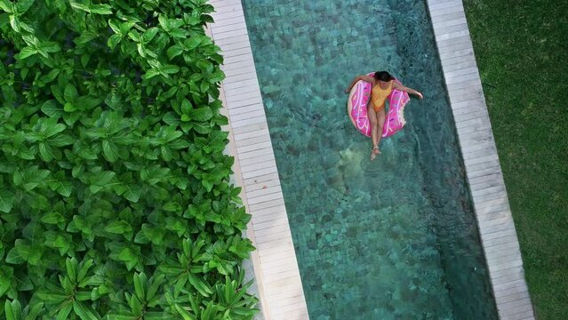 Top view of young woman in swimsuit on the pink donut lilo in the swimming pool. Female relaxing on inflatable float the pool at villa. Inflatable ring and mattress. Summer holiday idyllic. High view