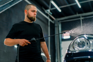 Fototapeta na wymiar A male car wash employee applies car wash detergent to a black luxury car using a spray gun in the car wash box