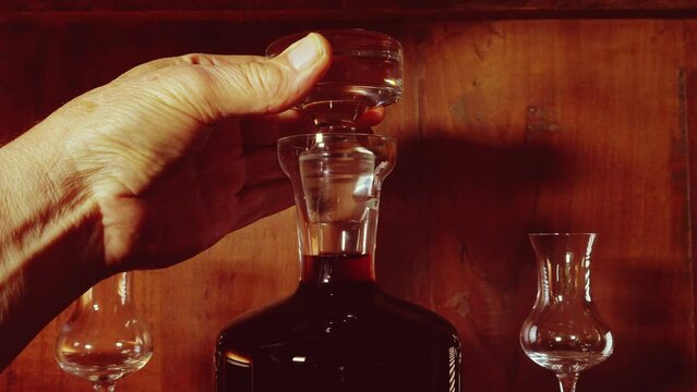 Closeup of man&rsquo;s hand putting the stopper head into a drinks decanter full of red wine, sherry or port, with two glasses nearby, on a shelf in a wooden cabinet or cupboard.