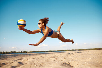 Dynamic image of young woman in motion, playing beach volleyball, hitting ball and falling down on...