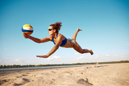Dynamic image of young woman in motion, playing beach volleyball, hitting ball and falling down on sand. Sunny warm day. Concept of sport, active and healthy lifestyle, hobby, summertime, ad